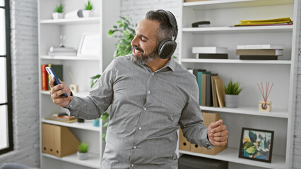 A mature hispanic man with a beard and grey hair enjoys music while dancing in an office with a smartphone and headphones.