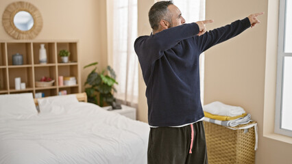 A mature hispanic man with grey hair and beard stretches in a well-lit, cozy bedroom interior,...