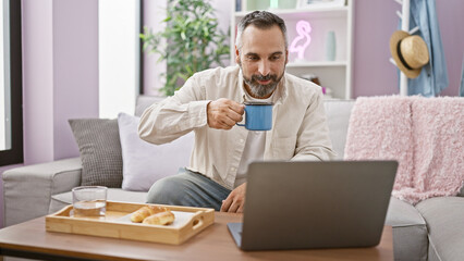 Mature hispanic man with beard using laptop and drinking coffee at home