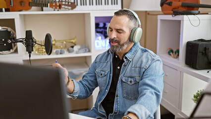 A content hispanic senior man with a beard wearing headphones enjoys music in an indoor studio...