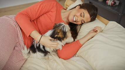 Smiling woman playing with biewer terrier on a cozy bed in a warmly lit bedroom.