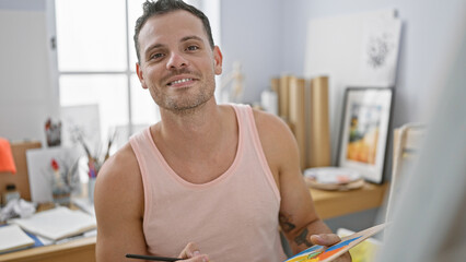 A smiling young hispanic man with a beard, wearing a tank top, holds a paintbrush in an art studio...