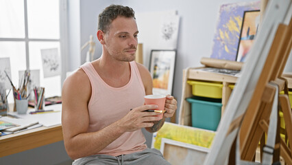 Handsome hispanic man holding coffee in an art studio, surrounded by paintings and bright interior.