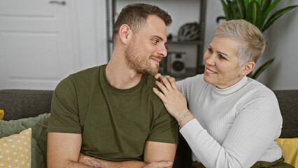 A mature woman and a younger man share a tender moment on a couch, indoors, suggesting a family bond.