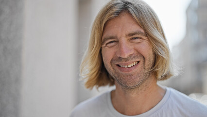 Handsome young adult blond man with long hair smiling in an urban outdoor street setting.