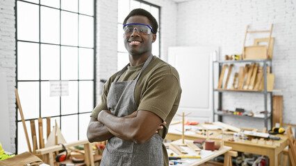 Confident african american man with arms crossed wearing safety glasses stands in a well-equipped carpentry workshop.