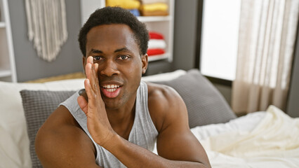 A smiling black man makes a secret-telling gesture in a cozy bedroom setting.