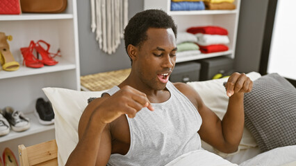 Young black man in tank top pointing down while sitting on bed indoors with shelves and shoes in background