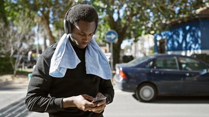 A young african american man with headphones uses a smartphone on a sunny urban street with cars in...