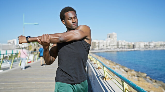 African american man stretching at a sunny seaside promenade with a clear blue ocean backdrop. - Powered by Adobe