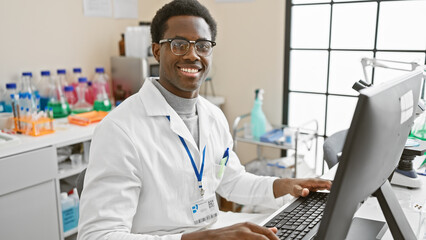 Smiling african american man wearing lab coat working on computer in clinical laboratory