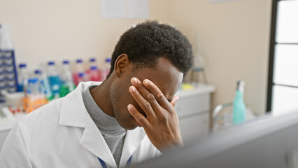Tired african american man in a white lab coat resting head indoors at a medical laboratory.