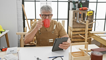 A grey-haired man enjoys a coffee break in a woodworking workshop, browsing on a tablet amidst handcrafted woodwork.
