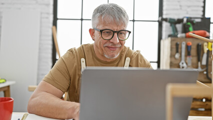 A smiling, mature man works attentively on a laptop in a sunny, tool-filled carpentry workshop.
