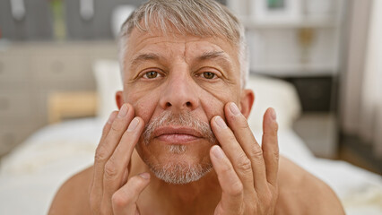 Close-up of a contemplative mature man with grey hair in a bedroom, scrutinizing his facial...