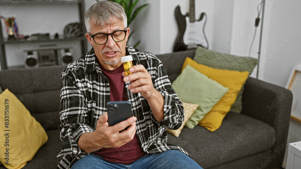 Sticker Mature man examines medication while holding phone in a cozy living room, concerned and perplexed.