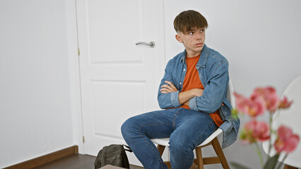 A pensive teen boy sits arms crossed in a minimalist white interior, exuding a moody vibe.