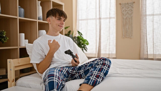 Blond teenager boy smiling while holding a video game controller, sitting on a bed in a cozy bedroom setting with a white shirt and plaid pajama pants.