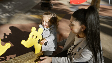 A smiling mother and daughter enjoying sunny day at an outdoor playground, bonding over a seesaw...