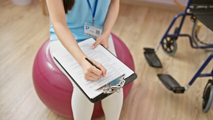 A young asian woman healthcare professional writes on a clipboard while sitting on a balance ball in a clinic.