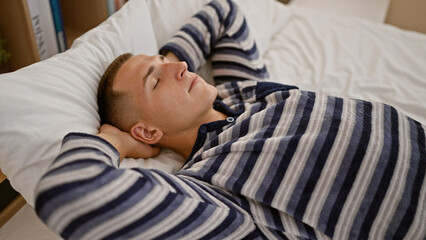 A young, handsome man in a striped shirt relaxes, eyes closed, in a comfortable bedroom setting.