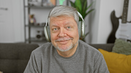 A senior man with headphones smiles warmly in a comfortably furnished living room, embodying leisure and relaxation at home.