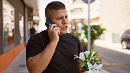 Handsome young latin guy engrossed in a serious talk over the phone, holding a colorful bouquet of flowers, standing casually under the sunny street.