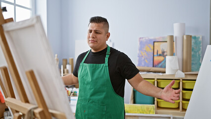 Upset young latin man artist, standing tiredly by his unfinished painting at the art studio