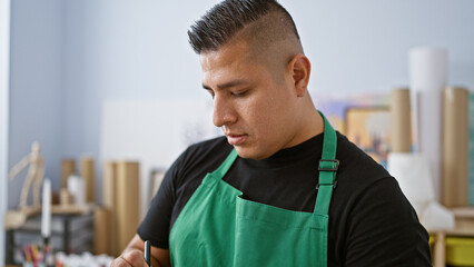 Handsome young latin man, an artist fully concentrated, standing with serious face indoors by his canvas, holding a paintbrush at art studio, focused on painting