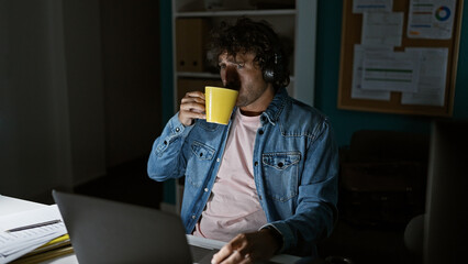 A hispanic man in a denim jacket sips from a mug while working late on a laptop in a dim office...