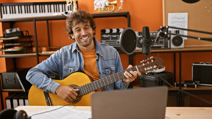 Smiling hispanic man playing guitar in a home music studio with recording equipment
