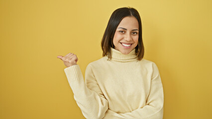 Smiling young hispanic woman pointing sideways against an isolated yellow background, portraying positivity and guidance.