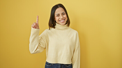 Smiling young hispanic woman in a cream sweater pointing upwards against a yellow background, exuding confidence and cheerfulness.
