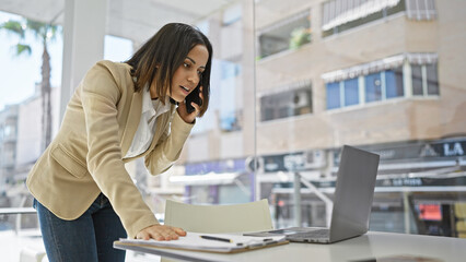 A professional hispanic woman in a beige suit uses a smartphone and laptop in a modern office with a cityscape view.