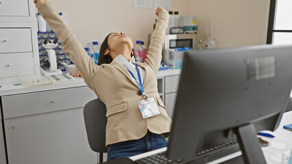 A content woman scientist stretches joyfully indoors at her laboratory workstation, reflecting relaxation and job satisfaction.
