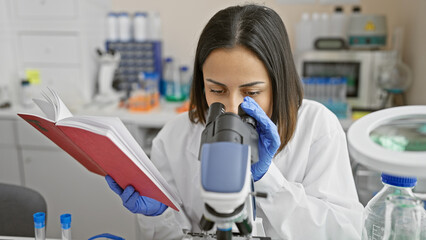A focused woman scientist analyzing specimens under a microscope while holding a research journal...
