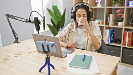 Hispanic woman drinks water while podcasting in a studio with laptop and microphone setup, recorded...