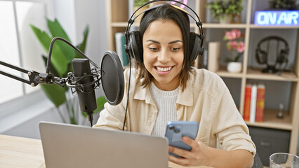 Smiling young woman with headphones in a radio studio holding a smartphone while recording a...