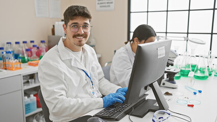 In a bright laboratory, a woman and a man in lab coats work on a computer, surrounded by scientific equipment.