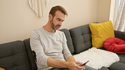 Middle-aged man with beard sitting on couch using smartphone in cozy living room