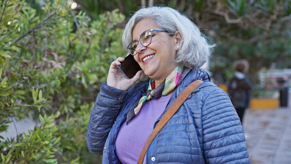 Cheerful mature woman with grey hair talking on smartphone in a city park, surrounded by greenery.