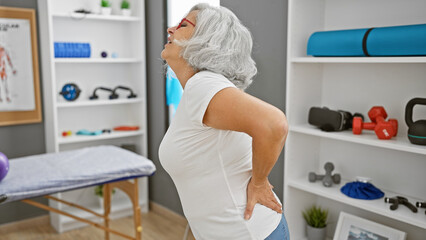 An elderly woman stands in pain inside a rehabilitation clinic, hinting at a possible back problem.