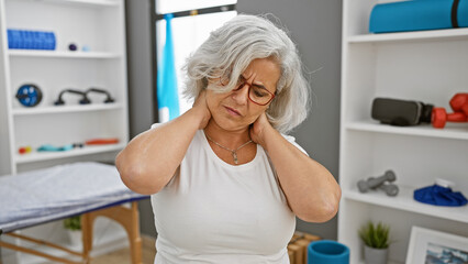 Mature woman in pain holding neck indoors at a rehabilitation clinic, with medical equipment in the background.