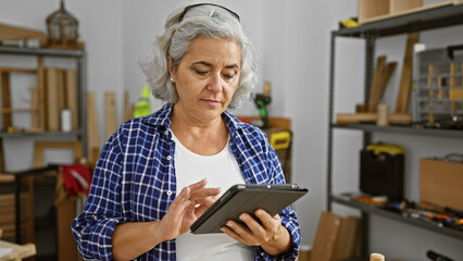 A mature woman uses a tablet in a well-organized carpentry workshop, surrounded by wooden shelves and tools.