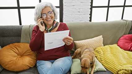 A smiling woman talks on the phone while holding a document, with a dog beside her on a couch in a...