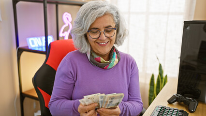 A smiling woman counts polish zloty bills indoors, near a gaming setup with a neon sign and plant.