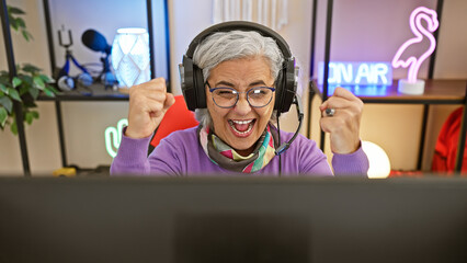 Enthusiastic elderly woman with headphones in a gaming room celebrating a victory at night.