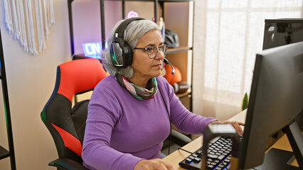 Mature woman gaming indoors with headphones looking at computer screen in a well-lit room