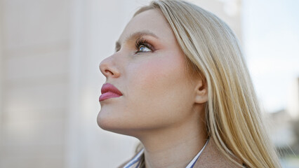 A contemplative young blonde woman gazes upward on an urban street.