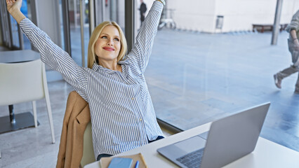 Relaxed blonde woman stretching at a modern office with city reflection on window, showcasing urban work lifestyle.